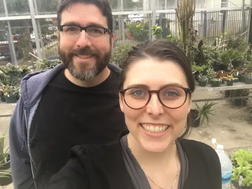 A man and a woman smiling at the camera, in a greenhouse