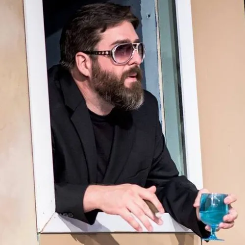 Man in sunglasses holding a cigar and a drink and leaning out of a window